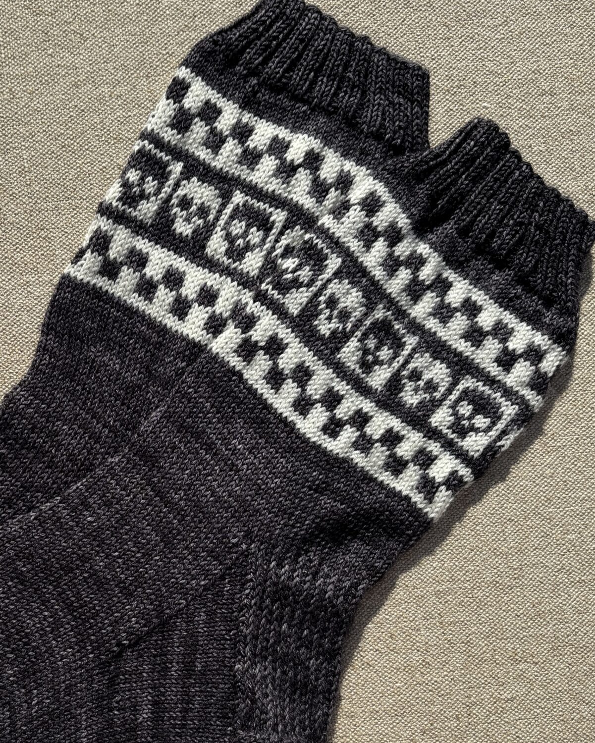 Close-up of a thick, knitted scarf in black and gray with a light gray geometric stripe pattern laid over a beige fabric background.